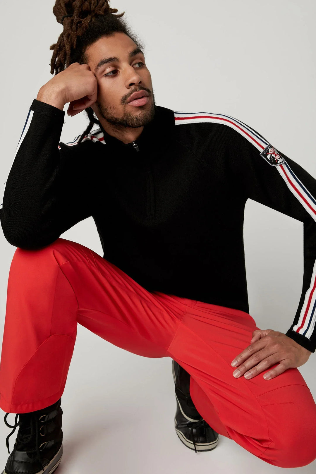 Man wearing red Davos ski pants and a black Chase men’s sweater by Fera paired with ski boots for his day on the slopes.