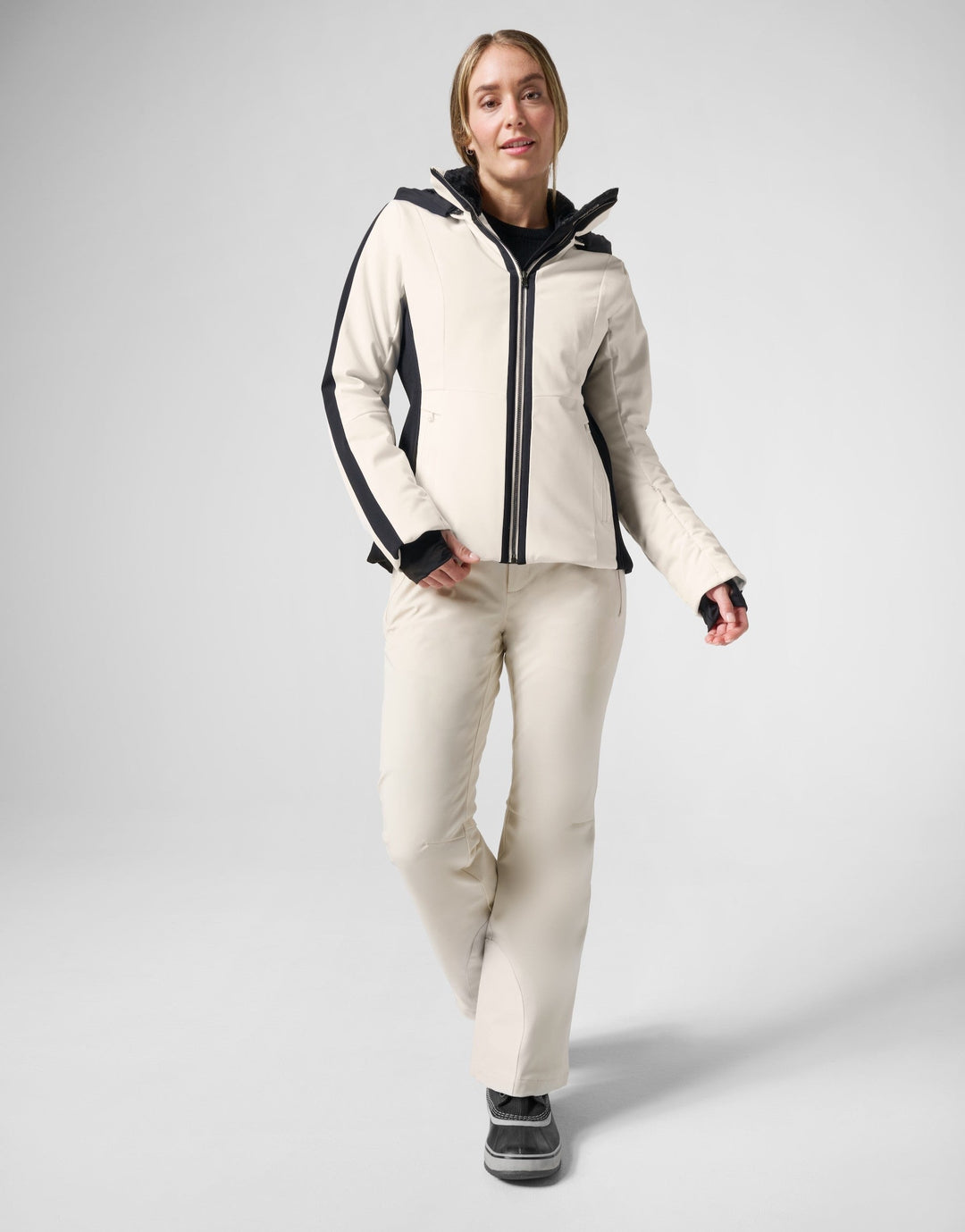 A woman wearing FERA ski pants and a jacket in cream with black accents, against a grey background.